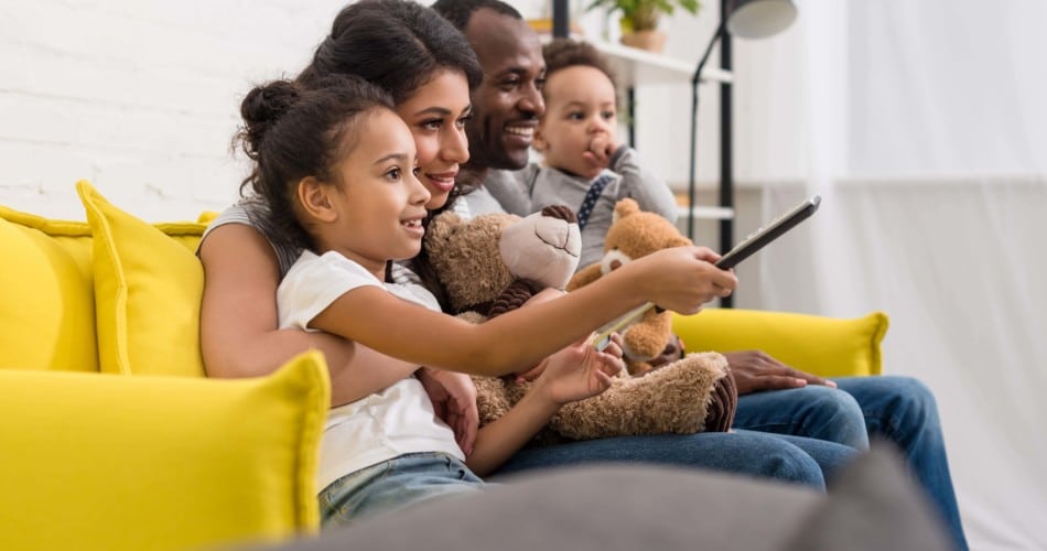 Medienkonsum: Eine vierköpfige Familie sitzt gemeinsam glücklich auf der Couch und schaut offensichtlich TV.