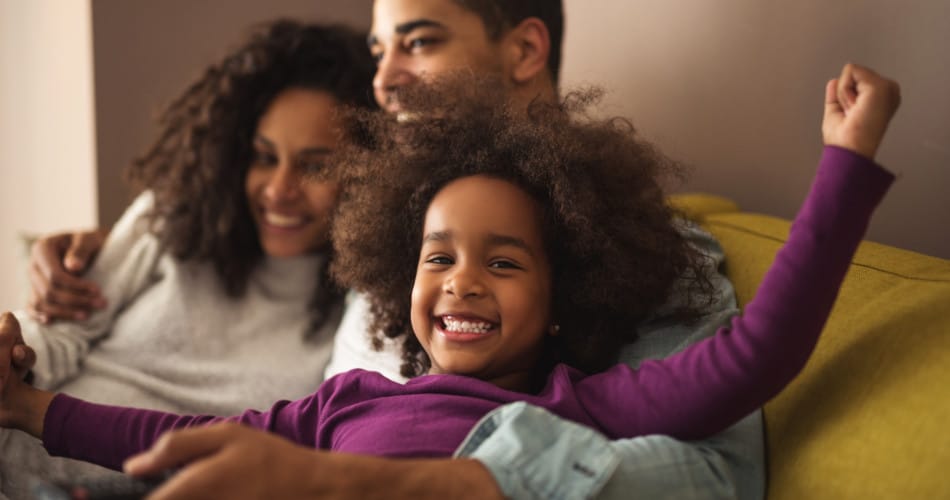 Eine afroamerikanische Familie bestehend aus Mutter, Papa und kleiner Tochter verbringt zu Hause auf der Couch Zeit miteinander. Der Vater hat eine Fernbedienung in der Hand.