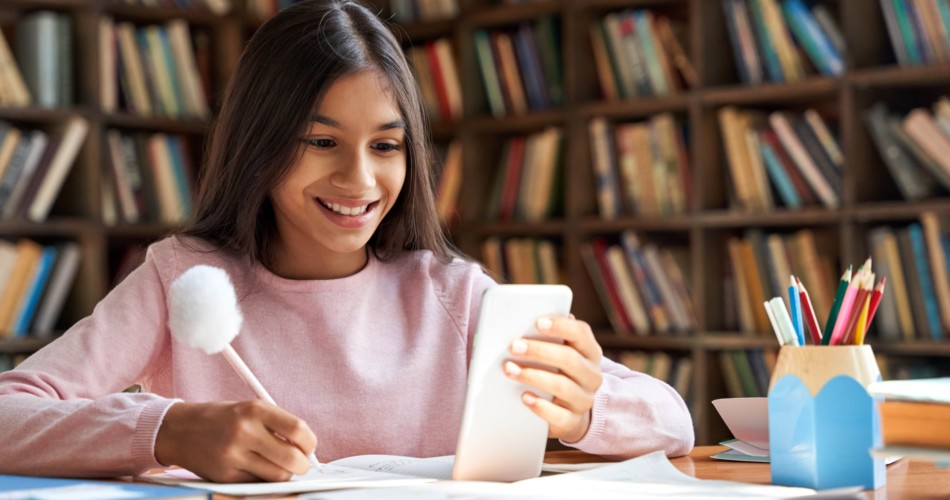 Lern-Apps für Kinder: Auf dem Bild sieht man ein junges Mädchen, dass am Schreibtisch sitzt und lernt. Sie schaut auf ihr Handy und schreibt dabei etwas auf. Sie lächelt dabei.