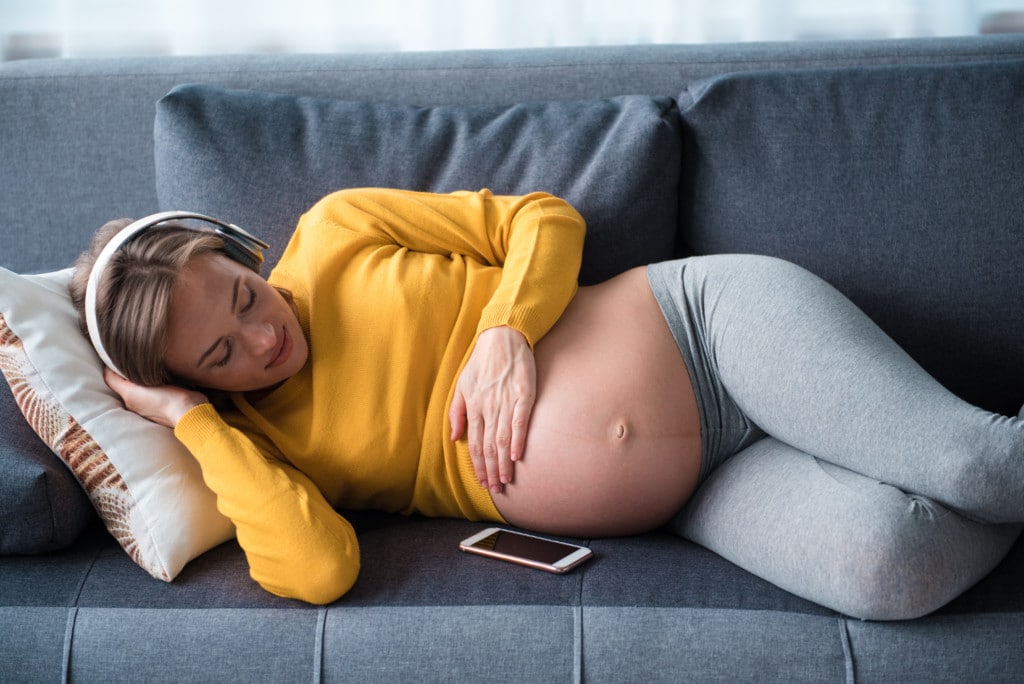 Eine schwangere Frau liegt beruhigt auf der Couch und hört Musik.