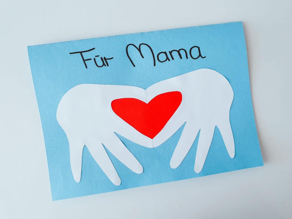 Eine hellblaue Karte, auf der Kinderhände ein Herz formen als Geschenk für Mama basteln.