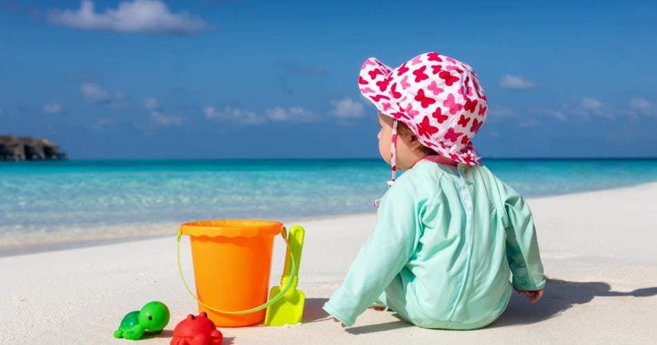 Ein Baby sitzt mit Kleidung und Hut am Strand - Titelbild für Sonnenschutz für Babys.