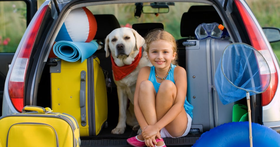 Auf dem BIld sieht man ein kleines Mädchen gemeinsam mit einem Hund. Beide sitzen im offenen Kofferraum und freuen sich auf die Autoreise.