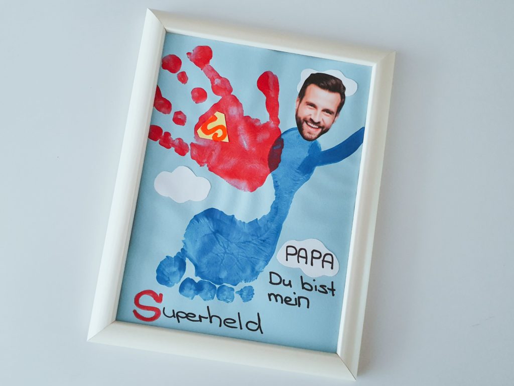 Ein Superheld-Bild aus dem Handabdruck und Fußabdruck eines Kindes und mit einem Foto des Papas als Kopf als Vatertagsgeschenk gebastelt.
