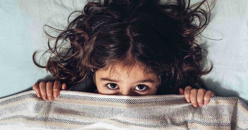 Ein kleines Mädchen liegt beängstigt im Bett und hält die Decke schützend vor ihr Gesicht.