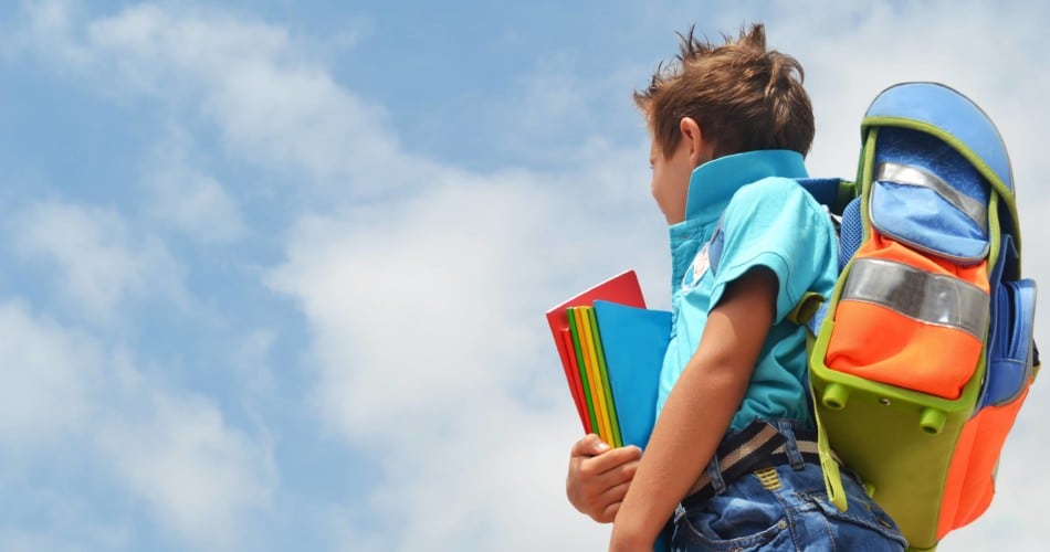 Ein Kind (Erstklässler) trägt einen Schulranzen auf dem Rücken und Schulmappen in seinen Hännden. Dabei schaut er in den Himmel.