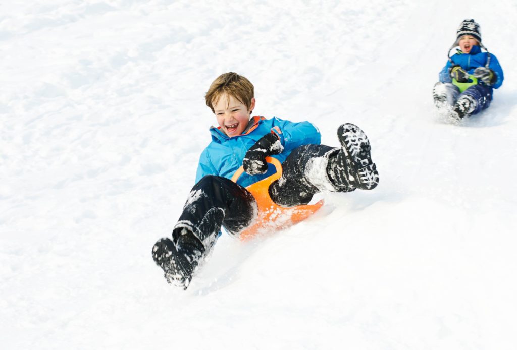 Zwei Kinder fahren mit Schlitten im Schnee in der Adventszeit einen Berg herunter.
