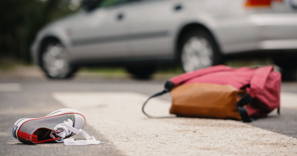 Ein Schuh und der Rucksack eines Schulkindes liegen auf der Straße. Im Hintergrund ein fahrendes Fahrzeug.